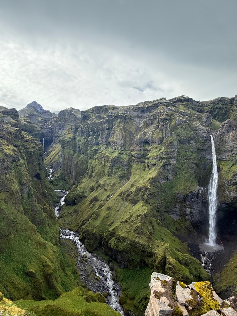two waterfalls within green cliffs or mountains coming together within a canyon with a river through it. 